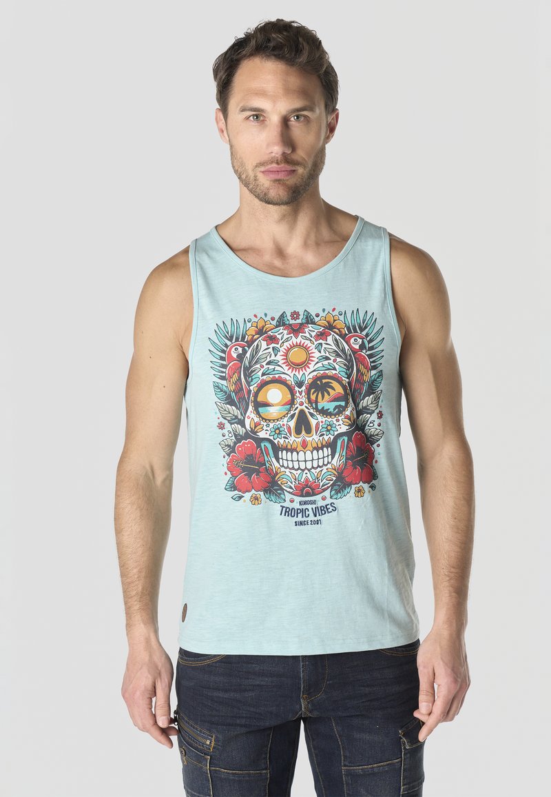 Hombre con camiseta sin mangas azul claro con diseño colorido de calavera tropical y flores, combinado con jeans azul oscuro.