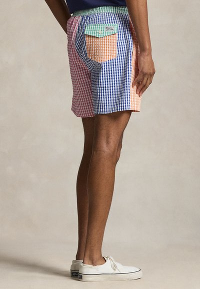 Άνδρας που φορά πολύχρωμα καρό σορτς με τσέπη και λευκά αθλητικά παπούτσια, στάσιμος πλάγια σε ουδέτερο φόντο.