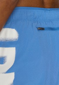 Calções de desporto azuis em tecido de malha texturizada, com um bolso com zíper e grandes letras brancas. O cós elástico proporciona flexibilidade.