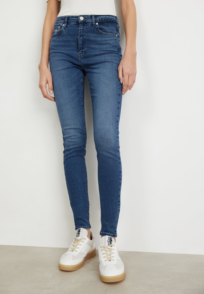 Jeans ajustados de denim azul medio, con una cintura alta, bolsillos cosidos y un ligero desvanecimiento. Combinados con zapatillas blancas.