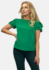 Zielona bluzka z krótkim rękawem, z falbaniastymi akcentami, miękka w dotyku, okrągły dekolt i luźny krój, zestawiona z ciemnymi spodniami.