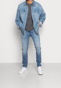 Ljusblå denimjacka med två bröstfickor, grå t-shirt, ljusblå jeans och vita sneakers. Jackan har en avslappnad passform.