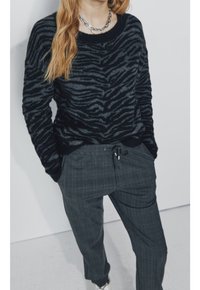 Pull rayé noir et gris avec un col rond, associé à un pantalon gris à carreaux. Le pull présente une texture douce et une coupe décontractée.
