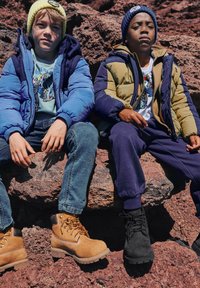 Dois rapazes sentados em rochas, vestidos com casacos acolchoados de cores vivas, jeans e botas. Um casaco é azul, o outro é verde e amarelo.