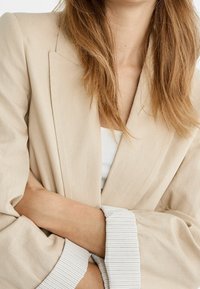 Blazer de lino beige con mangas anchas; los puños presentan una tela a rayas blanca y azul marino. Blusa blanca lisa debajo.