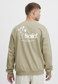 Tan mikina s bílou grafikou květiny v květináči, s logem "!solid" a textem "Nářadí a potřeby pro květinářství a zahradnictví" na zadní straně.