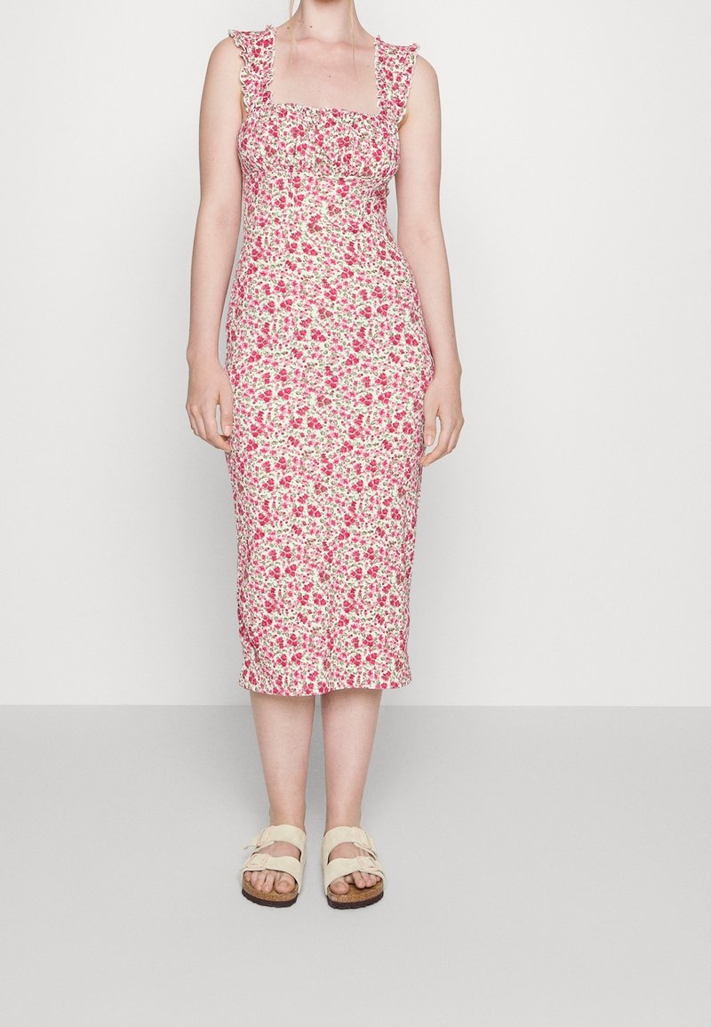 Femme portant une robe midi sans manches à encolure carrée, rose et blanche à motif floral, avec des bretelles à volants et des sandales beiges à boucle, debout devant un fond uni.