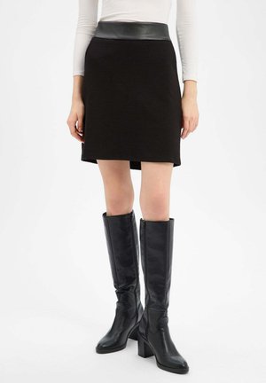 Frau trägt einen schwarzen, strukturierten Rock mit einem Lederbund und schwarzen kniehohen Stiefeln mit Absatz, stehend vor einem schlichten weißen Hintergrund.