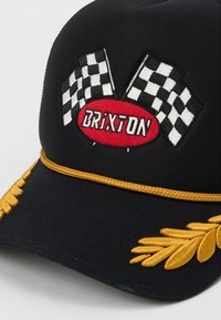 Czarna czapka z haftem w szachownicę, czerwonym akcentem z logo "Brixton" oraz detalem ze złotego haftu wzdłuż daszka. Płaski daszek.