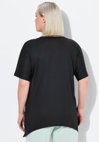 Černé tričko s krátkým rukávem uvolněného střihu, hladkého materiálu a zakřiveným lemem, nošené přes světlé kalhoty. Žádné viditelné logo.