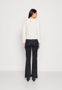 Balta adīta blūze ar teksturētu rakstu un pūkainām piedurknēm, apvienota ar tumšiem platiem džinsiem. Aizmugurē ir neliels atslēgas caurums.