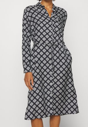 Sort knapkjole med hvidt blomsterprint, lange ærmer, krave, taljebånd og sidelommer båret af en person.