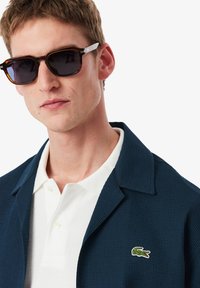 Ναυτικό υφασμάτινο σακάκι πάνω από λευκό πουκάμισο με κολάρο, με μικρό πράσινο λογότυπο. Μαύρα ορθογώνια γυαλιά ηλίου με καφέ αποχρώσεις.