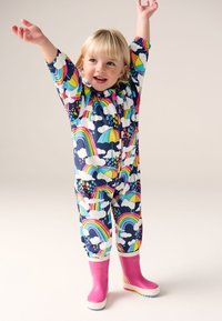 Colorful rain suit featuring a navy background with rainbow, umbrella, and cloud patterns. Paired with pink rubber boots and white soles.