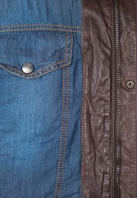 Džínová bunda s levým náprsním kapsou, kombinující modrý denim a hnědou kůži. Hnědé prošívání zvýrazňuje modrou látku a detaily zipu.