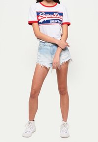 Weißes T-Shirt mit rotem Kragen und Ärmeln, mit blauem und rotem Logo-Text. Getragen mit hellblauen, ausgefransten Jeansshorts und weißen Sneakern.