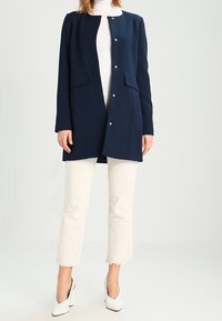 Cappotto blu navy, lungo fino al ginocchio, con scollo rotondo, chiusure a pressione, due tasche frontali e texture liscia, abbinato a jeans bianchi e tacchi.