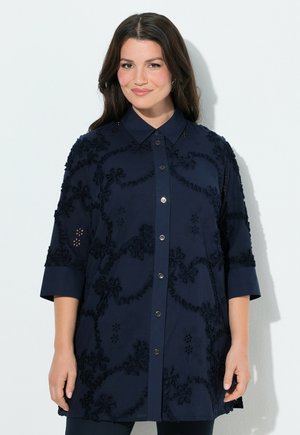 Lächelnde Frau mit langen dunklen Haaren, die ein marineblaues, geknöpftes Hemdblusenkleid mit Blumenstickerei und 3/4-Ärmeln trägt.