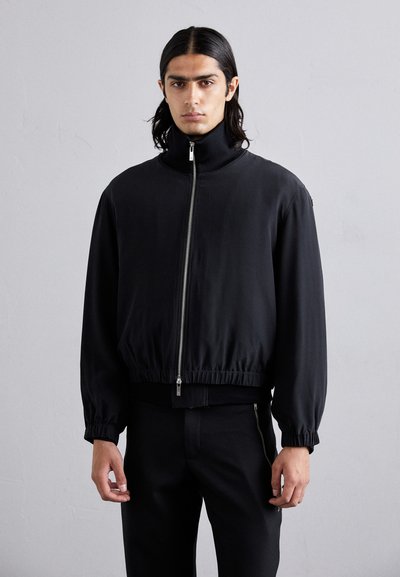 Schwarze Bomberjacke mit hohem Kragen, elastischen Bündchen und Saum sowie einem Frontreißverschluss. Glattes Material mit matter Oberfläche.