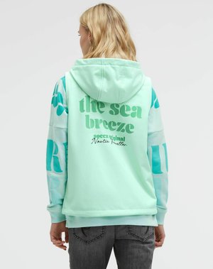 Blonde person wearing mint green hoodie with teal text "the sea breeze" and blue patterned sleeves, facing away on plain background.