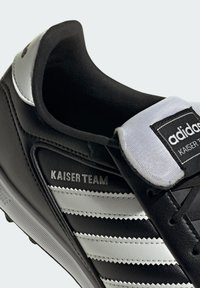 Sneaker Adidas Kaiser Team nera con strisce bianche, etichetta sulla linguetta bianca e dettaglio argento sul tallone, mostrata in primo piano.