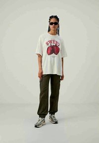 Άτομο που φοράει γυαλιά ηλίου, υπερμεγέθη λευκή μπλούζα με μεγάλες κόκκινες φράουλες και κείμενο "SWEET", σκούρα cargo παντελόνια και γκρι αθλητικά παπούτσια.