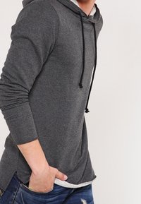 Felpa grigia con maniche lunghe, cappuccio con cordino e fessure laterali. Realizzata in tessuto morbido, presenta un orlo interno testurizzato. Indossata con jeans blu.