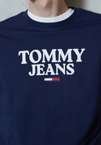 Námořnická modrá bavlněná tričko s kulatým výstřihem, s velkým bílým textem "TOMMY JEANS" a horizontálním akcentem červené, bílé a modré pruhy níže.