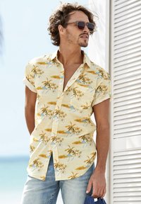 Camicia a maniche corte con bottoni di colore giallo chiaro, decorata con un motivo di spiagge e palme. Il tessuto sembra leggero e liscio.