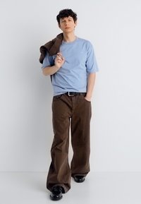 Νέος άνδρας που φοράει ανοιχτό μπλε μπλουζάκι και φαρδιά καφέ παντελόνια, κρατώντας ένα καφέ σακάκι πάνω στον ώμο του, στέκεται μπροστά σε ένα απλό λευκό φόντο.