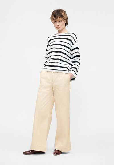 Νεαρό άτομο με κοντά μαλλιά, φορούσε μακρυμάνικο πουλόβερ με λευκές και μαύρες ρίγες, μπεζ παντελόνι φαρδύ, και καφέ παπούτσια, στέκεται με τα χέρια στις τσέπες.