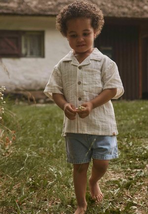 Junges Kind mit lockigem Haar, trägt ein beiges Shirt und Jeansshorts, läuft barfuß auf Gras und hält kleine weiße Blumen.