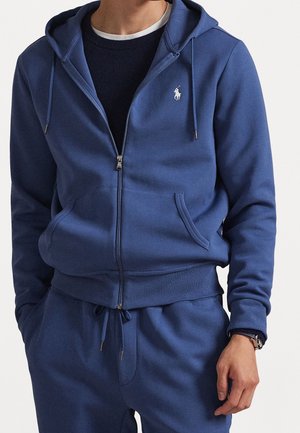 Mann trägt einen blauen Kapuzenpullover mit Reißverschluss und Taschen sowie passende blaue Jogginghosen, ein schwarzes Shirt darunter und eine Armbanduhr am linken Handgelenk.