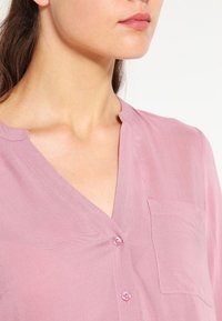 Blouse rose clair en tissu transparent avec un décolleté en V, des manches longues, une petite poche et des boutons sur le devant.