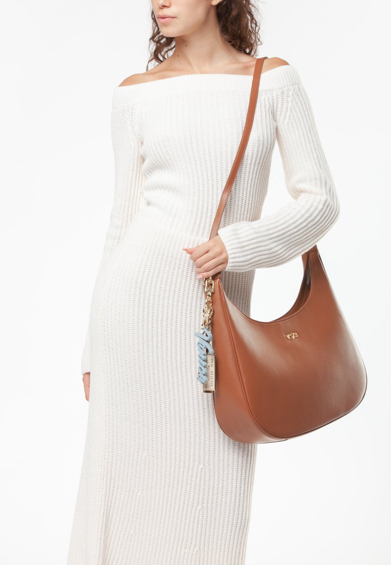 Bolso de hombro de cuero marrón con una textura suave y forma curved; presenta un charm azul y herrajes dorados. Llevado con un vestido largo blanco de canalé.