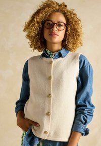 Hellbeige Wolle-Weste mit goldenen Knöpfen über einem Jeanshemd. Lockige Haare und ein gemusterter Schal sind sichtbar. Einfaches Design und neutrale Farben.