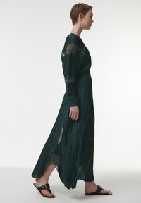 Largo vestido plisado en verde oscuro con mangas translúcidas y texturizadas, y una cintura ajustada. Combinado con sandalias planas negras con una hebilla dorada.