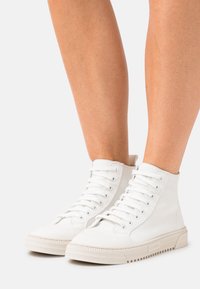 Jambes portant des baskets montantes en toile blanche à lacets, debout sur un fond gris clair.