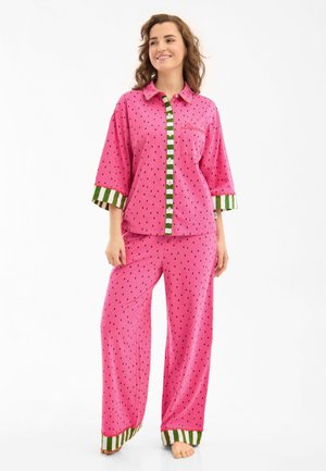 Woman wearing loose pink pajama set with black seed-like dots, green and white striped accents on cuffs, collar, and button placket.
