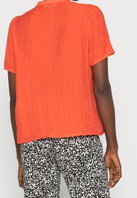 Short-sleeve, textured orange top with a relaxed fit, displayed with patterned black and white pants featuring an abstract design.