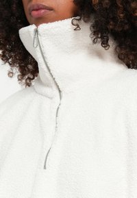 Witte fleece pullover met hoge kraag en halve ritssluiting, voorzien van een zachte, textuurafwerking en subtiele zilveren hardwaredetails.