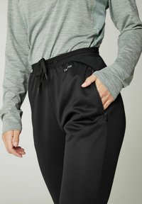 Zwarte sportieve broek met een gladde textuur, voorzien van een tailleband met trekkoord, een zijzak met rits en een logo-detail. Gecombineerd met een grijze lange mouwtop met patroon.