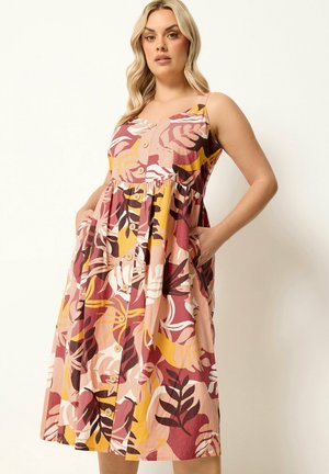 Donna che indossa un vestito midi senza maniche con bottoni sul davanti e motivo multicolore di foglie tropicali, che posa con le mani nelle tasche.