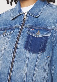 Džínová bunda s světle modrým odstínem, vybavená tmavě modrou náprsní kapsou, kovovým zipem a dvěma předními klopovými kapsami. Texturovaná tkanina.