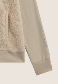 Felpa beige con un pannello di pelliccia morbida e testurizzata sulla parte anteriore e "REBOOK TRAINING" in strass sulla manica.