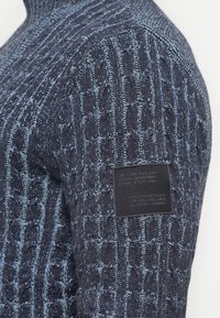 Gebreide trui in donkerblauw met lichtblauwe accenten, voorzien van een gestructureerd kabelpatroon. Inclusief een zwart merklabel op de mouw.