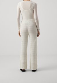 Bílý body s dlouhým rukávem a kulatým výstřihem na zádech v kombinaci s krémovými, texturovanými širokými kalhotami s vertikálními pruhy a zvlněným lemem.