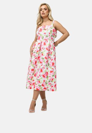 Vrouw draagt een mouwloze, wit midilengte jurk met roze en groene bloemenpatronen, poseert met één hand op haar heup en zilveren hakken.