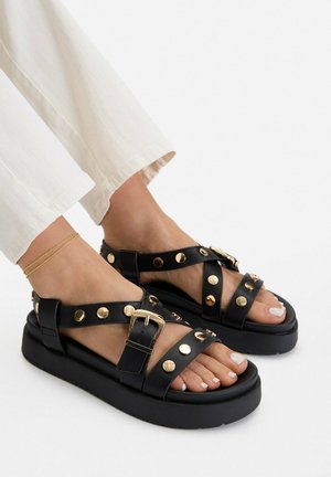 Füße in schwarzen Plateausandalen mit goldenen Nieten und Schnallen, kombiniert mit weißen Hosen und einem dünnen goldenen Fußkettchen.