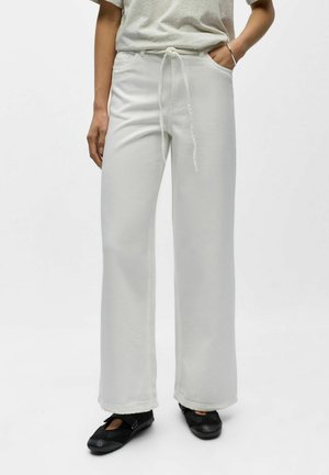 Person iført løstsittende hvite bukser med snor og svarte slip-on sko, stående mot en ensfarget lys bakgrunn.